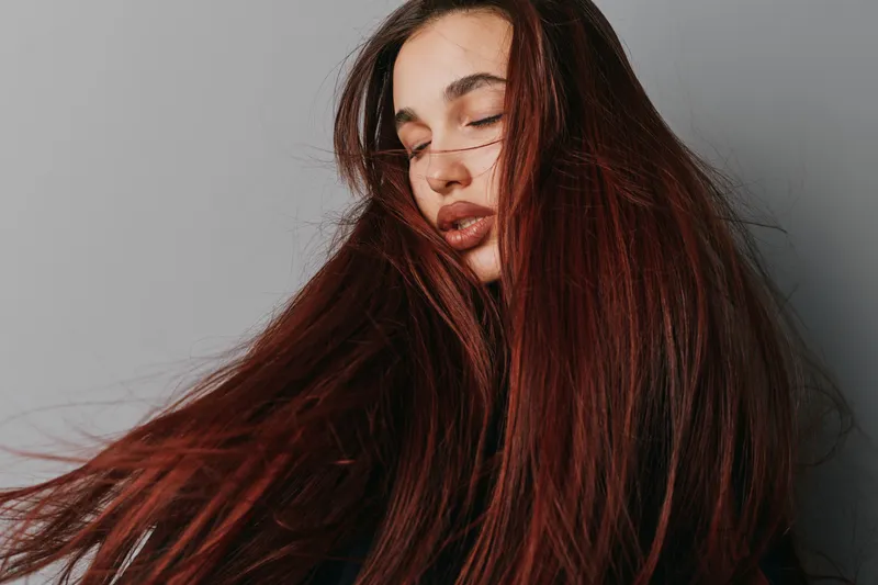 Woman with long red hair and closed eyes posing on gray background. Female model with smooth skin and natural makeup in studio with flowing hair movement and calm expression.