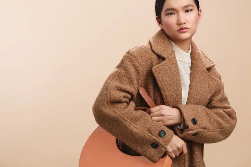 Asian woman wearing a brown coat holds a guitar case in a neutral studio, presenting a confident and poised demeanor with a subtle smile.