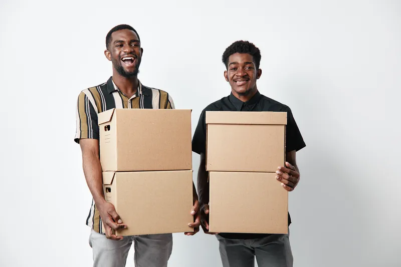 Two African American men with dark skin holding cardboard boxes for delivery and moving, smiling and standing on white background in casual clothing for logistics and shipping.