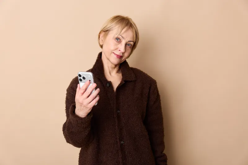 Woman smartphone smiling communication technology casual portrait studio beige background, she holds a phone for messages, warm light, close face, everyday lifestyle, relaxed mood