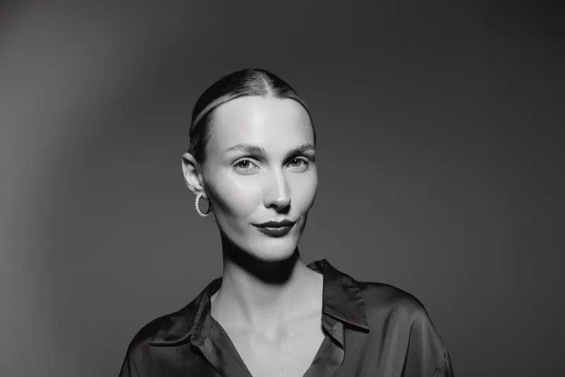 Portrait of a fair skinned Caucasian woman with slicked back hair and hoop earrings wearing a dark blouse. Studio lighting creates a dramatic, elegant mood for fashion or beauty.