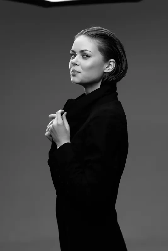 Black and white portrait of a confident woman in a dark coat with braided hair posing against a plain background.