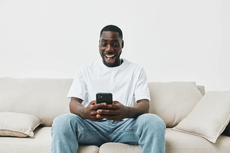 Happy young African man enjoying social media on smartphone while sitting on a beige couch at home, showcasing casual style and positive emotions