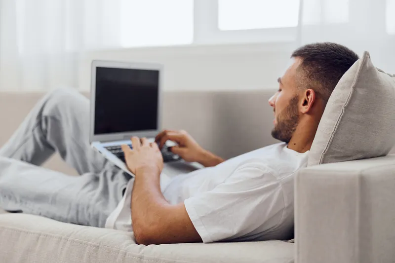 Relaxed man working on a laptop while lounging on a sofa, showcasing a modern home office vibe in a light, airy environment filled with natural light.