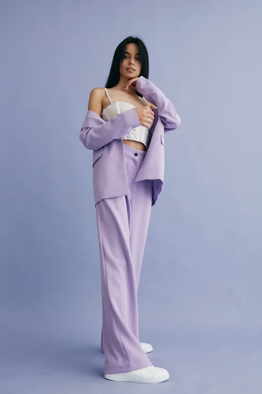 young woman in lilac suit and white top posing confidently against plain lavender background, showcasing stylish modern fashion and casual elegance.