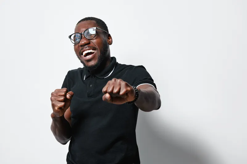 African American man with dark skin and glasses smiling and posing with fists up in black polo shirt on white background. Happy, confident and energetic male portrait.