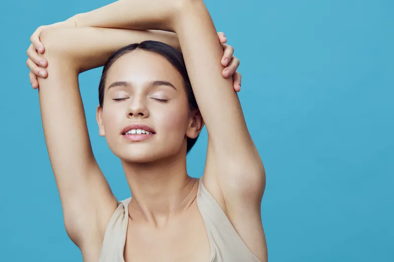 Young woman in a beige top stretching her arms with closed eyes, embracing relaxation against a solid blue background, conveying a sense of calm and tranquility