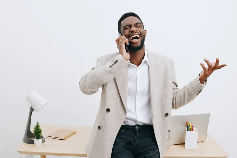 Confident smiling man talking on phone while standing in modern office with wooden desk, laptop, plant, and office supplies, dressed in smart casual attire, enjoying work moment, indoor workspace, bus