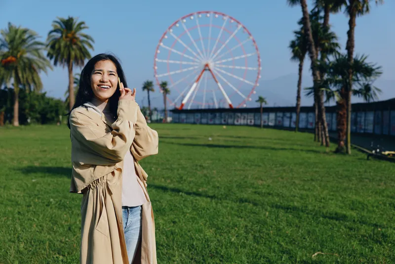 Smiling woman in a park foreground with a ferris wheel in the background on a sunny day, outdoor leisure scene with green grass and palm trees, capturing joy and travel vibes