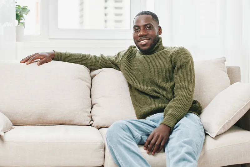 Smiling African man in a cozy green sweater, relaxing on a sofa at home, conveying warmth and comfort in a modern living space
