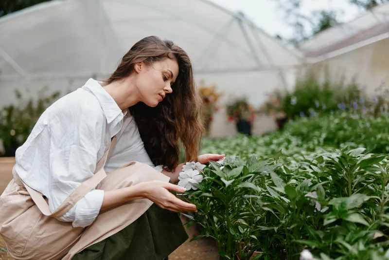 Beautiful woman in a greenhouse, gently touching flowers, showcasing the connection with nature, wearing a light outfit, amidst greenery and vibrant colors