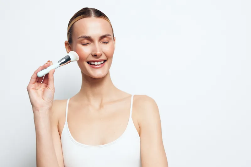Young woman with blonde hair applies makeup with a cosmetic brush on her cheek while smiling with closed eyes against a white background in a white tank top.