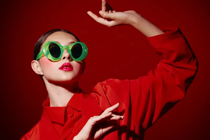 Fashion model wearing oversized green sunglasses and bold red lipstick poses against red background in a stylized beauty creative concept involving vivid colors and dramatic lighting.