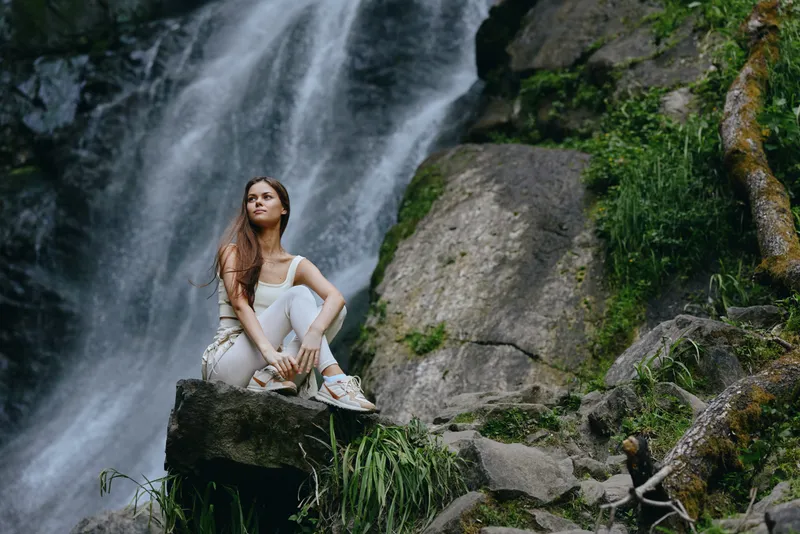 young woman sitting on rock near waterfall, wearing casual white clothes, surrounded by lush green nature, serene and contemplative outdoors scene