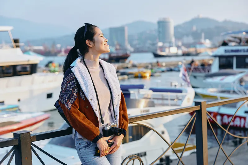Woman in a warm coat with scarf stands on a wooden pier beside a bustling harbor, boats in the background, clear sky and distant cityscape create a relaxed travel mood by the water