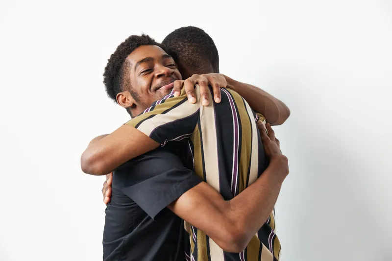 Two smiling black men sharing a warm embrace, showcasing friendship and connection. The colorful clothing adds vibrancy to the joyful atmosphere.