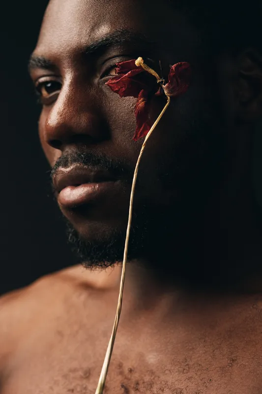 African man with dark skin holding dried red flower close to face in creative conceptual visual style portrait with moody lighting and artistic expression on black background
