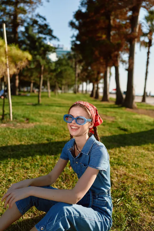 lifestyle woman in modern boho-western denim street style sitting on grass smiling with blue sunglasses and red bandana in warm film color sunlight