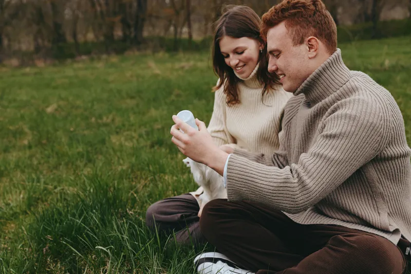 A young couple in warm sweaters sits on grass outdoors feeding a baby goat with a bottle during a relaxed lifestyle moment in nature.