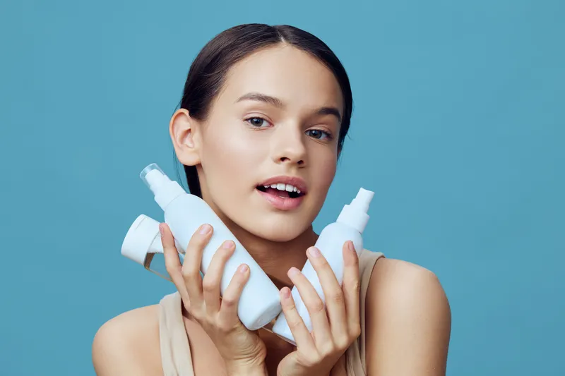 Young woman with skincare products in hands, showcasing beauty and wellness, against a light blue background, promoting healthy skin and self care routines