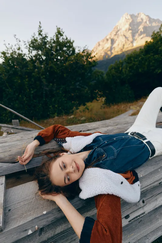 Woman mountain nature relaxation outdoors denim lying portrait travel leisure in a scenic alpine setting, young model reclining on a wooden bench by meadow and forest under warm sunlight.