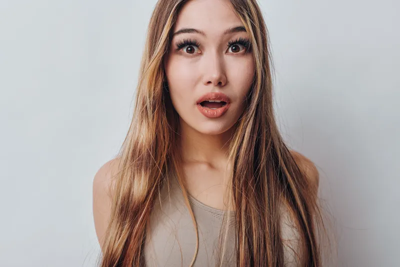 Young woman with long blonde hair looking surprised with wide eyes and open mouth in neutral studio background. Female emotion and expression closeup portrait.