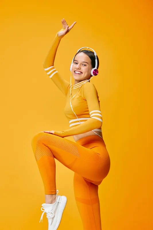 Vibrant studio scene with a woman in orange athletic wear dancing mid jump, wearing headphones, showcasing energy, fitness and sport in a bold playful pose against a bright orange backdrop.