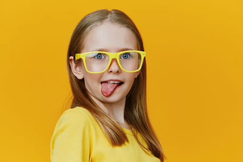 Cheerful girl with yellow glasses sticking out her tongue against a vibrant yellow background, showcasing fun expressions and playful fashion
