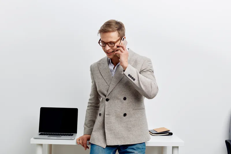 Businessman talking on mobile phone in modern office, casual grey blazer, glasses, smiling, professional, working, communication, technology, indoors, business environment, confident, male.