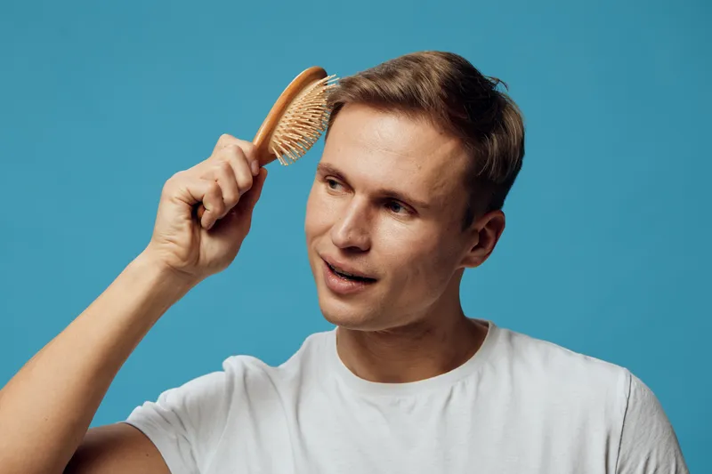 Man with short brown hair combing his hair with a wooden brush, wearing a casual white t-shirt, cheerful expression, isolated on blue background, studio portrait, lifestyle concept