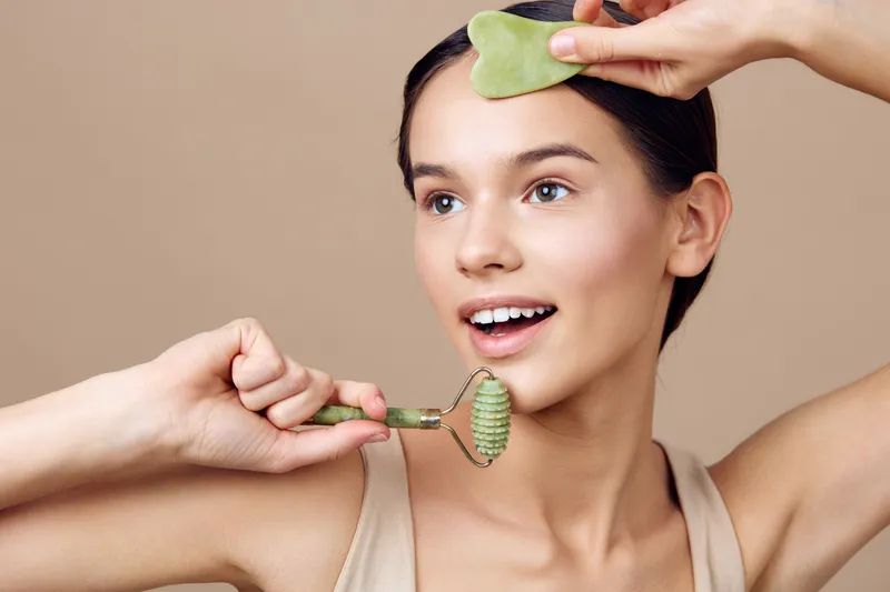 Young woman practicing jade rolling skincare routine with a gua sha stone, showcasing healthy skin and a joyful expression on a soft beige background