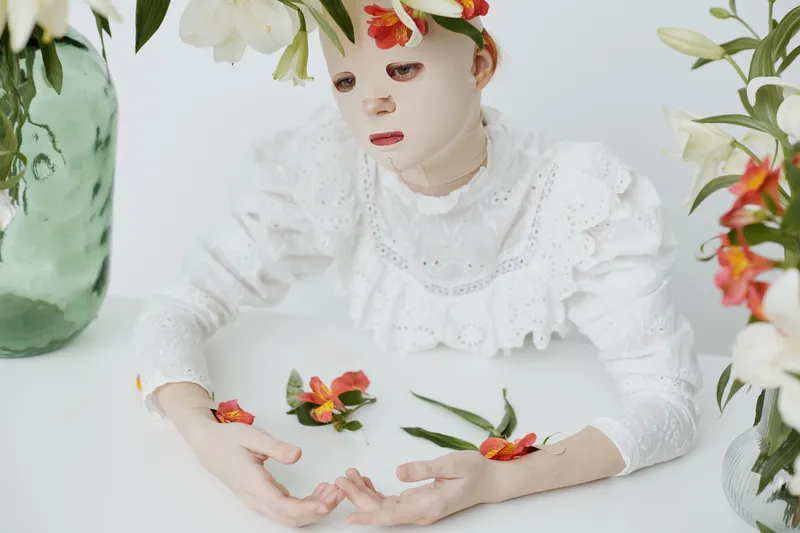 A serene model in a white ruffled dress wearing a facial mask surrounded by vibrant flowers, conveying emotions of tranquility and introspection in a minimalistic setting