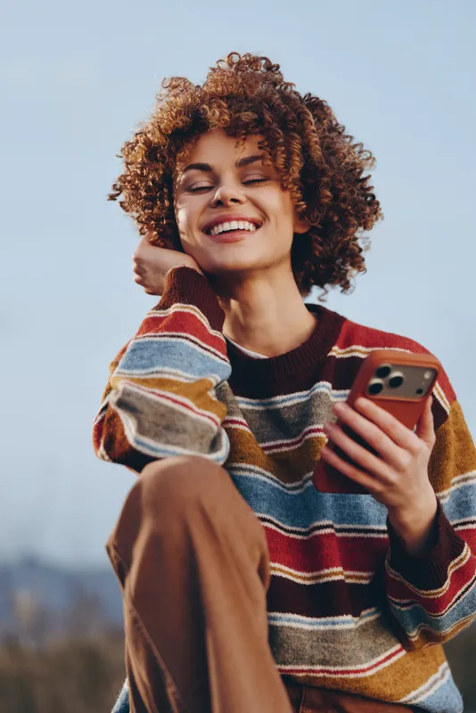 Woman with curly hair smiles, holding a smartphone outdoors, lifestyle concept, striped sweater, youthful relaxed portrait, open sky background, soft daylight