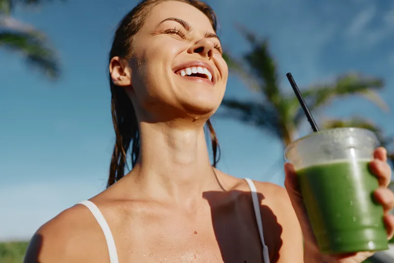 Smiling woman outdoors holds a green smoothie in a clear cup under bright sun, white tank top, blue sky, and palm trees creating a vibrant lifestyle scene focused on health and refreshment.