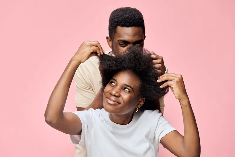 A joyful young African couple playfully styling their natural hair against a soft pink background, showcasing love, intimacy, and self expression through hair care