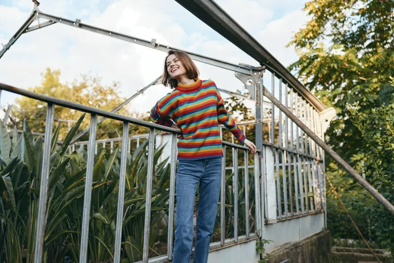 Woman in colorful striped sweater and jeans smiling in a greenhouse with lush plants, outdoor portrait of happy young adult leaning on metal railing for relaxed lifestyle scene