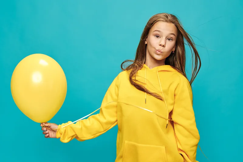 Girl with long hair in yellow hoodie holds yellow balloon on blue background. Casual portrait of child with playful expression and colorful outfit in studio setting.