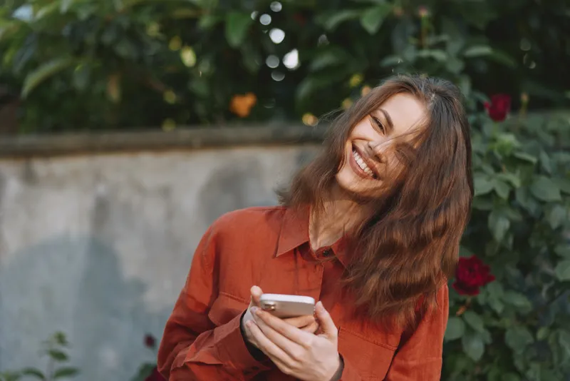 Woman lifestyle outdoors, smiling, red shirt, holding phone, sunlit garden background, casual moment, natural colors, greenery, breeze, joyful, relaxed