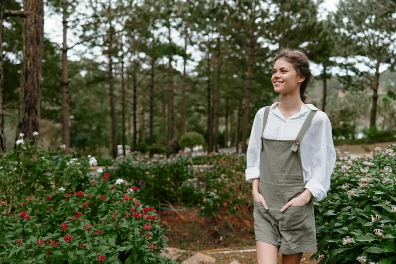 Smiling young woman wearing a casual outfit stands among blooming flowers in a serene garden, expressing joy and connection with nature