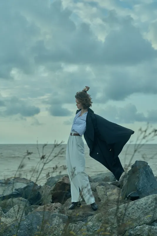 Windy coastal scene with a flowing cape and white attire on a rocky shore, moody clouds above, capturing freedom, fashion and adventure in a dramatic ocean setting