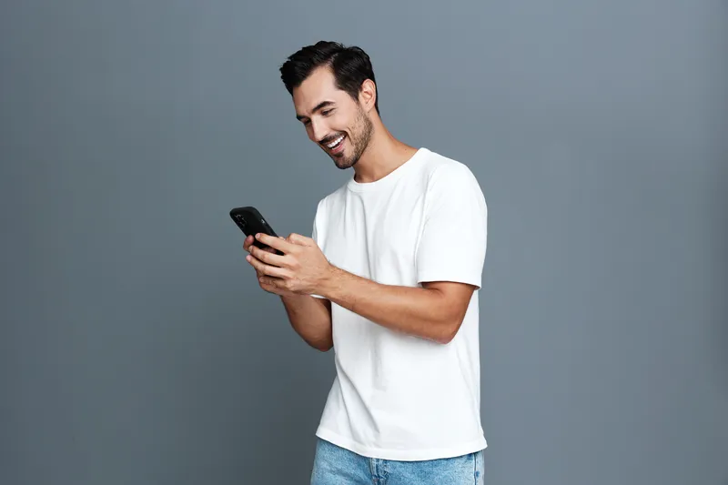 Happy smiling man using smartphone, wearing casual white t-shirt, standing against gray background, enjoying technology and modern lifestyle concept.