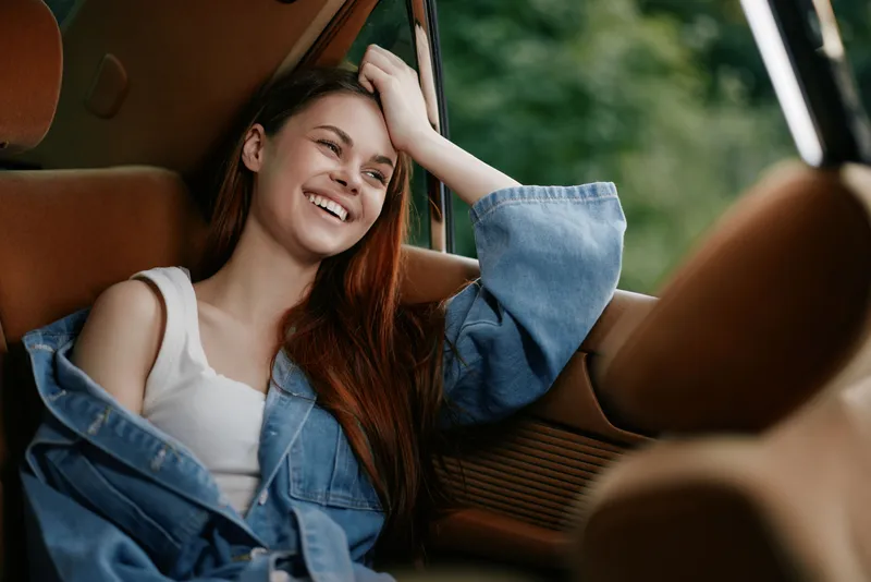 Young woman with long red hair smiling in a denim jacket sitting in a car, enjoying a sunny day outdoors, showcasing warmth and happiness.