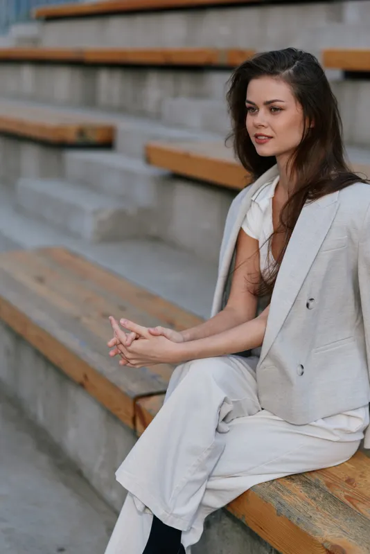 Woman in white stylish outfit sitting thoughtfully on wooden stairs with calm expression in outdoor urban setting, natural lighting, casual fashion concept