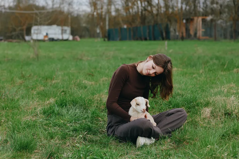 Cheerful woman outdoors, sitting on grass, gently cuddling a small white dog, soft sunlight, relaxed posture, gentle breeze, natural colors, serene countryside background on a sunny day with gentle