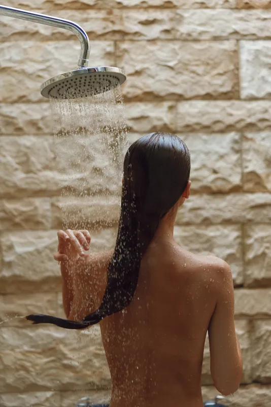 Woman showering with long wet hair under rain showerhead against textured beige stone tiles, natural skin, intimate moment of personal care and relaxation.