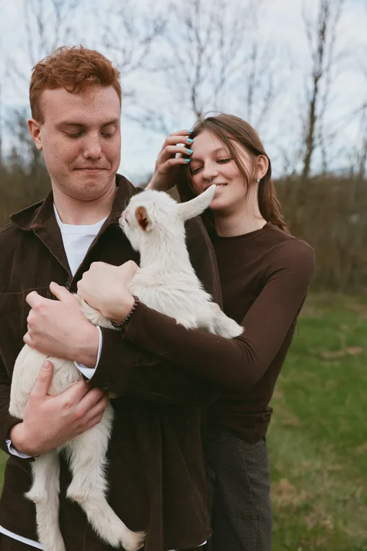 A couple hold a goat kid in a sunlit outdoors scene, sharing affectionate smiles and a gentle bond. A candid lifestyle image capturing natural happiness, kinship, and rural farm charm.