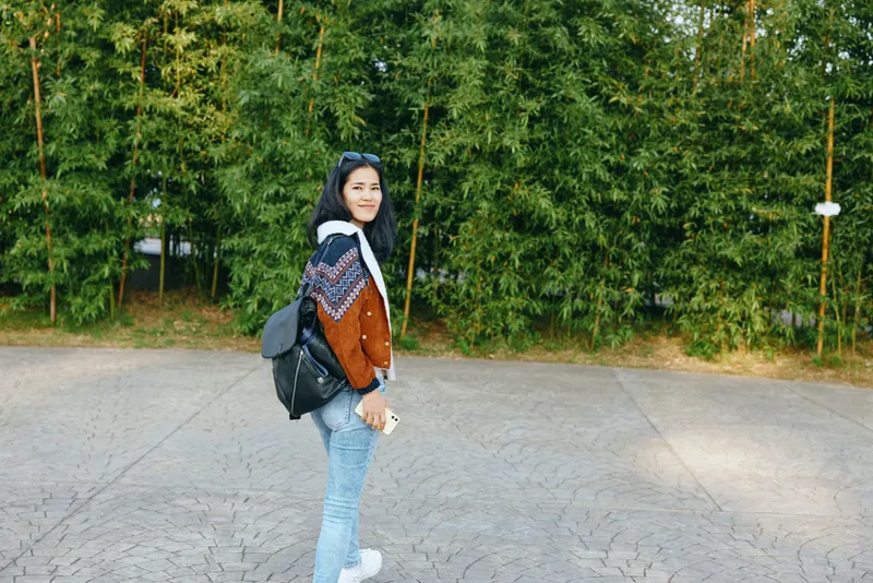 Woman with backpack walking along sunlit pavement beside trees in casual fashion, patterned sweater and jeans, sneakers and sunglasses, showcasing outdoor exploration and everyday street style daily