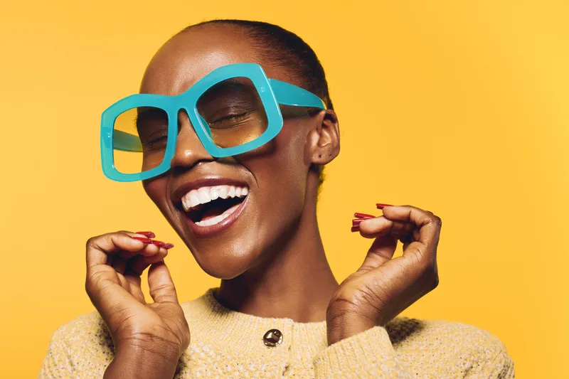 Happy African American woman with dark skin wearing large blue sunglasses and beige sweater smiles joyfully on yellow background. Concept of fashion, beauty, and positive emotions.