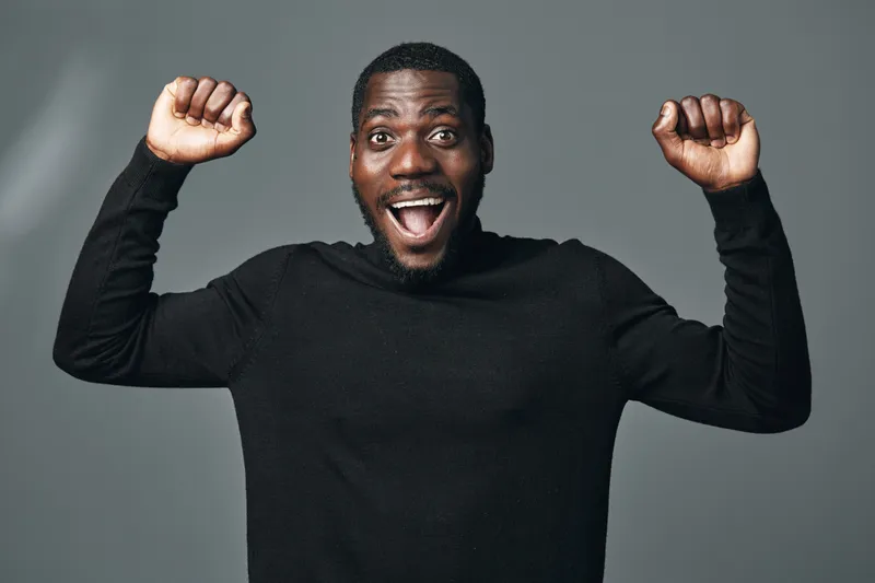 African man with dark skin wearing black sweater raising fists in excitement and joy in creative conceptual visual style isolated on gray background.