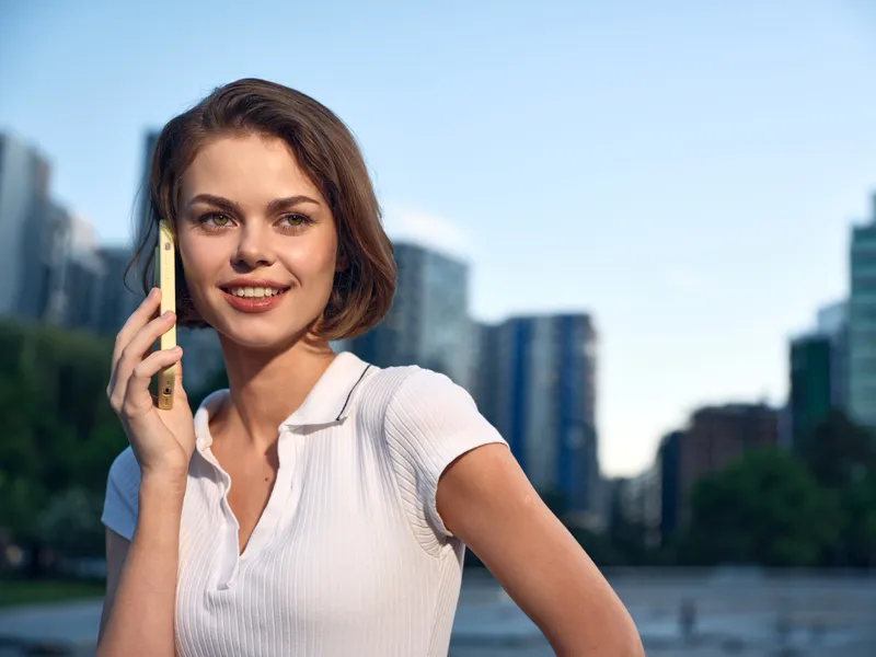 A cheerful young woman talks on a smartphone in a modern city setting. She smiles warmly, projecting confidence, connectivity, and a positive, upbeat mood in a bustling environment.